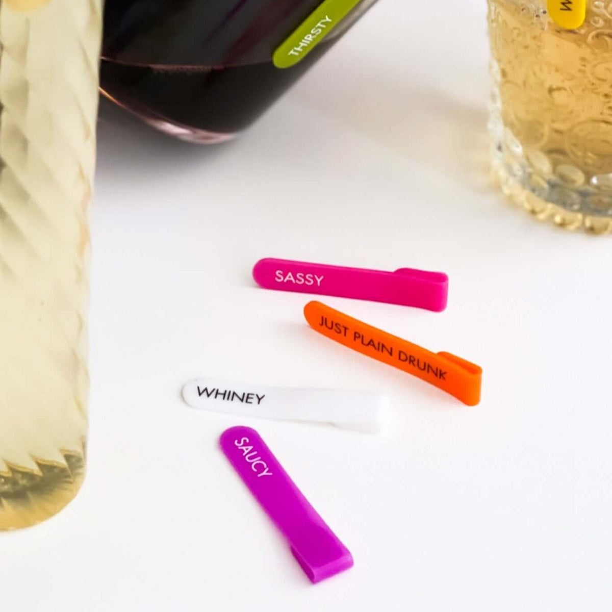 Colorful mini silicone identifiers for stemless glasses, featuring labels like 'Sassy' and 'Whiney', on a table.