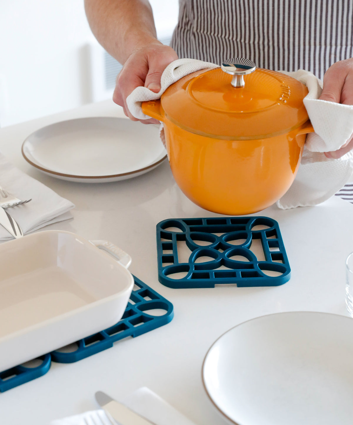 Person holding an orange pot over an interlocking silicone trivet on a dining table with plates.