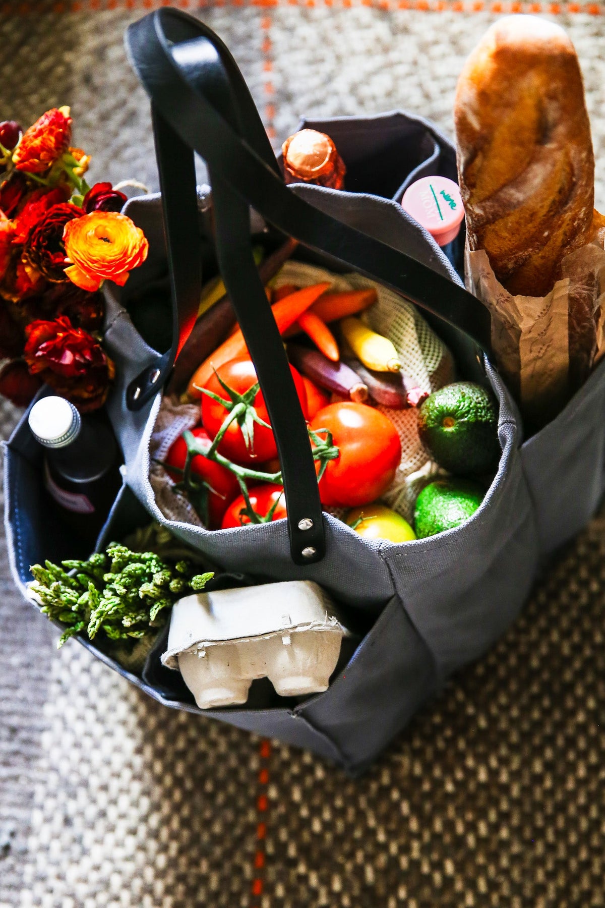 A grey multi-pocketed market tote filled with fresh vegetables, eggs, and a loaf of bread, ideal for grocery shopping.