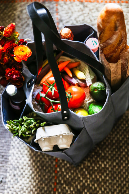 A grey multi-pocketed market tote filled with fresh vegetables, eggs, and a loaf of bread, ideal for grocery shopping.